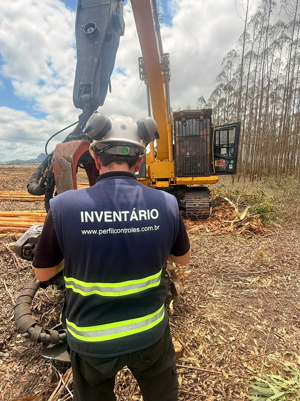 Técnico da Perfil Soluções, uniforme com identificação Inventário e www.perfilcontroles.com.br, de costas observando máquina florestal em campo, com toras empilhadas ao fundo.