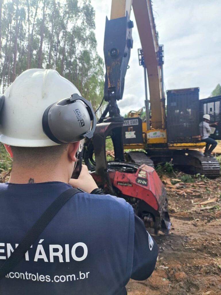 Técnico da Perfil registrando inventário de máquina florestal Harvester em campo, com equipe em plantação de eucalipto ao fundo.